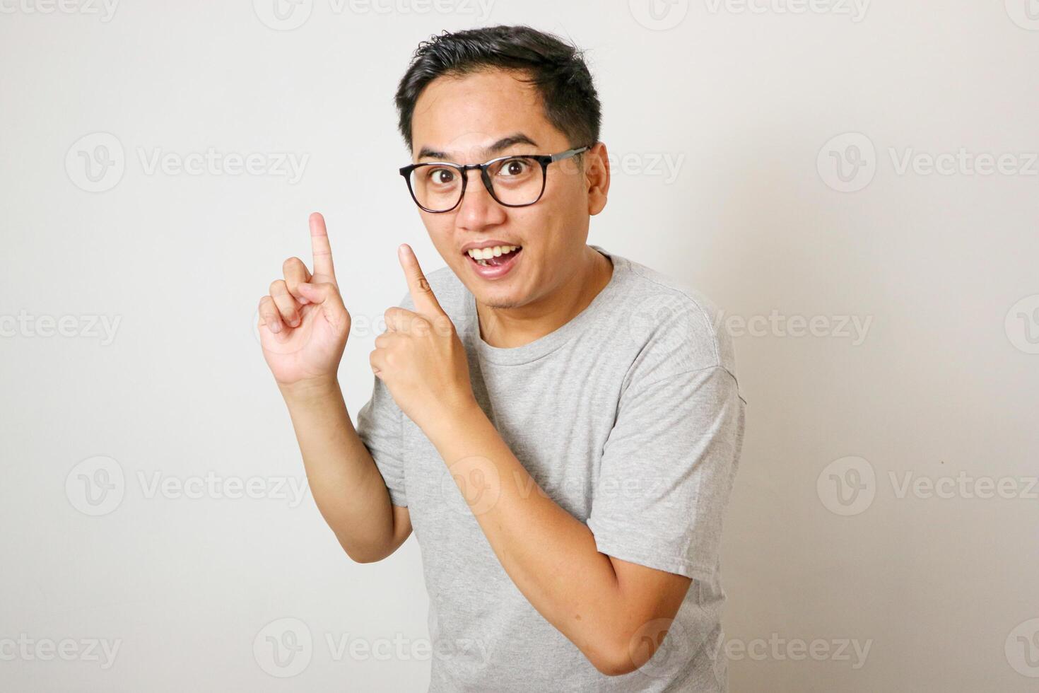 A young Asian man, wearing a grey shirt, looks thoughtfully upward while pointing with his right index finger. The plain white background highlights the subject's contemplative photo