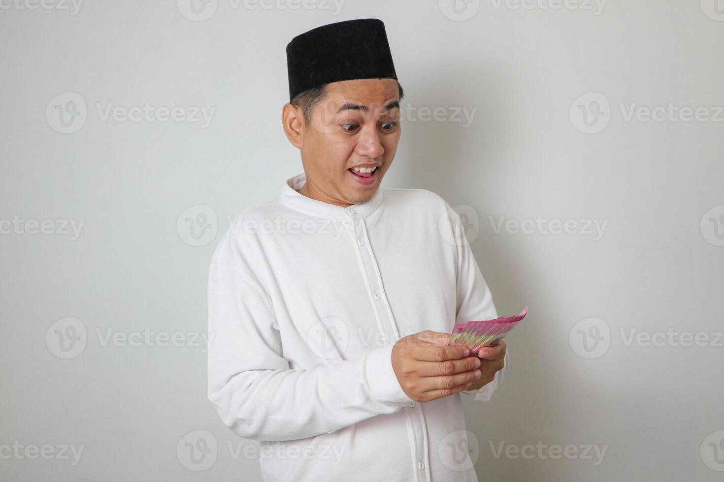 Excited young Asian Muslim man holding cash money rupiah banknotes and celebrating success isolated on white background. People religious Islamic lifestyle concept photo