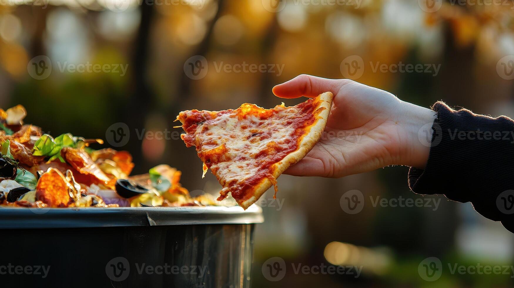 A hand reaches for a slice of pizza above a bin filled with discarded food. photo