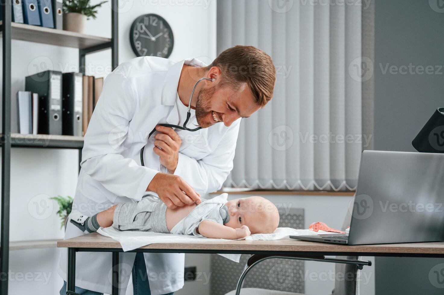 Using ,stethoscope.. Doctor in white coat is taking care of little toddler indoors photo