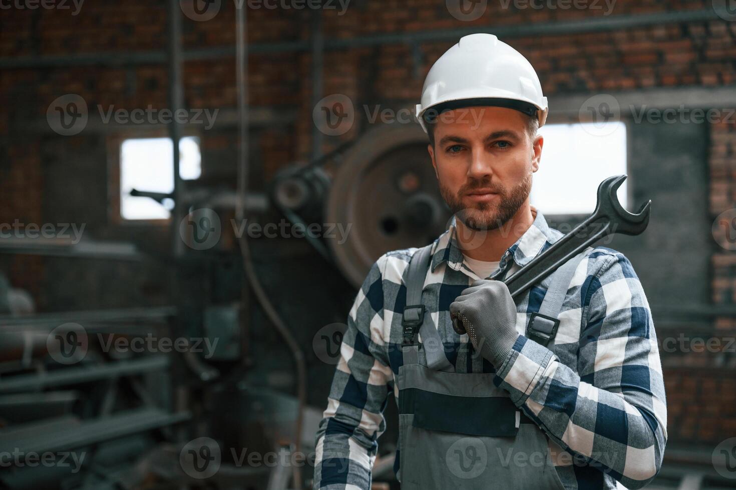 Standing in grey uniform and in white hard hat. Factory male worker is indoors photo