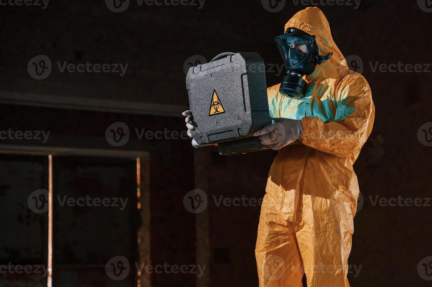 Blue light is going from ,hard case that is with hazard sign. Man dressed in chemical protection suit in the ruins of the post apocalyptic building photo
