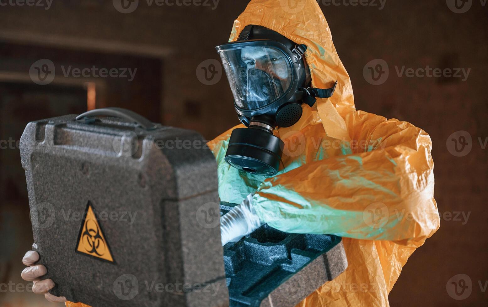 Blue light is going from hard case that is with hazard ,sign. Man dressed in chemical protection suit in the ruins of the post apocalyptic building photo