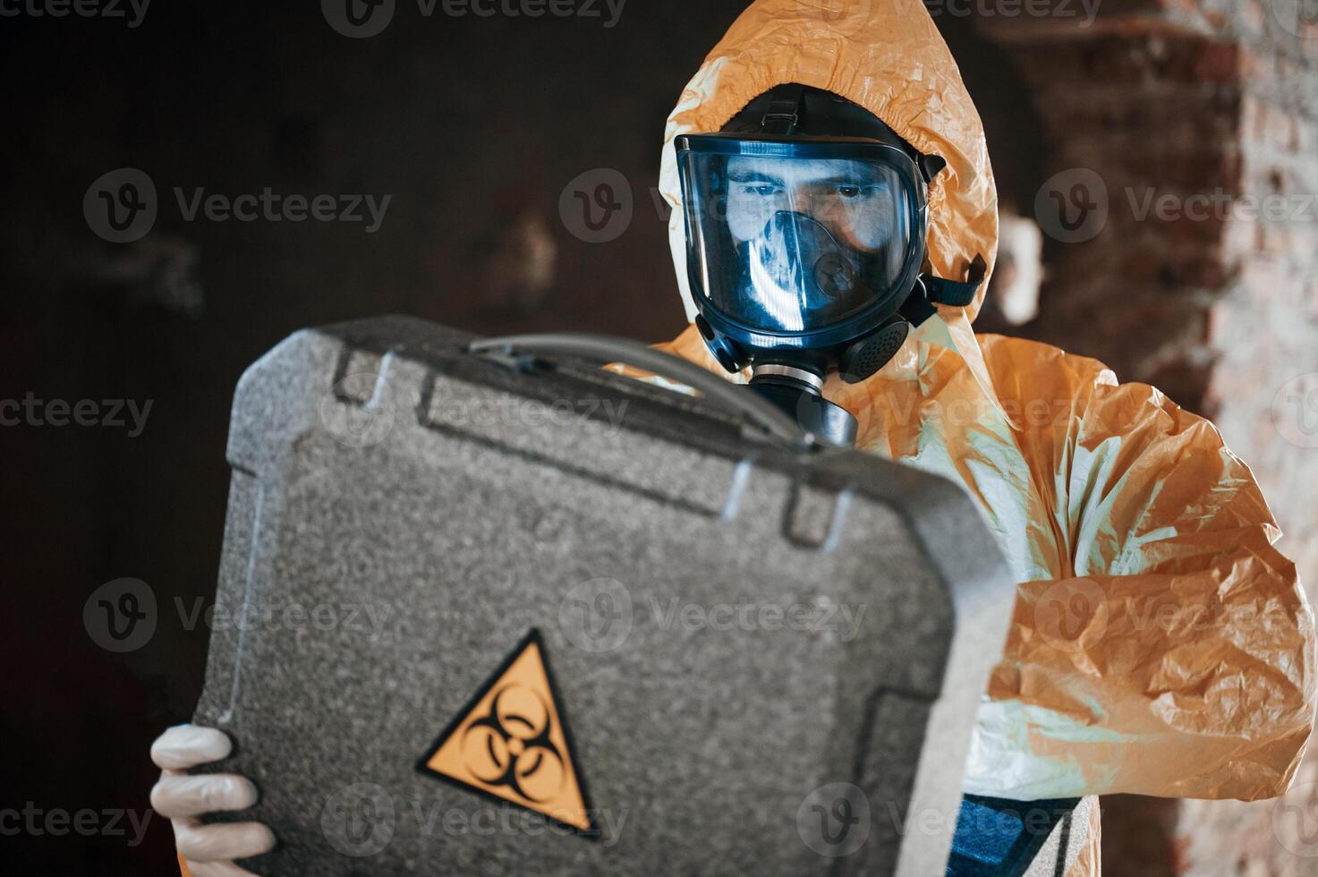 Blue light is going from hard case that is with hazard sign. Man dressed in chemical protection suit in the ruins of the post apocalyptic building photo