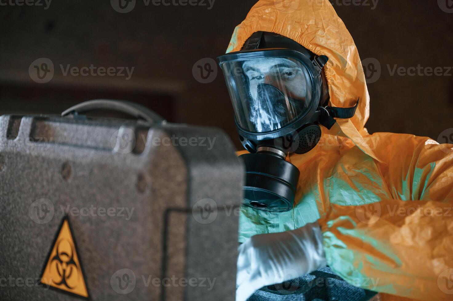 Blue light is going ,from hard case that is with hazard sign. Man dressed in chemical protection suit in the ruins of the post apocalyptic building photo