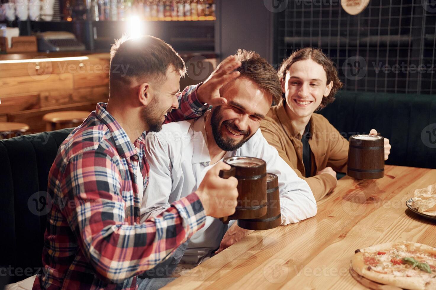 Smiling, knocking mugs, pizza on the table. Three friends are in the bar drinking beer and having a good time photo