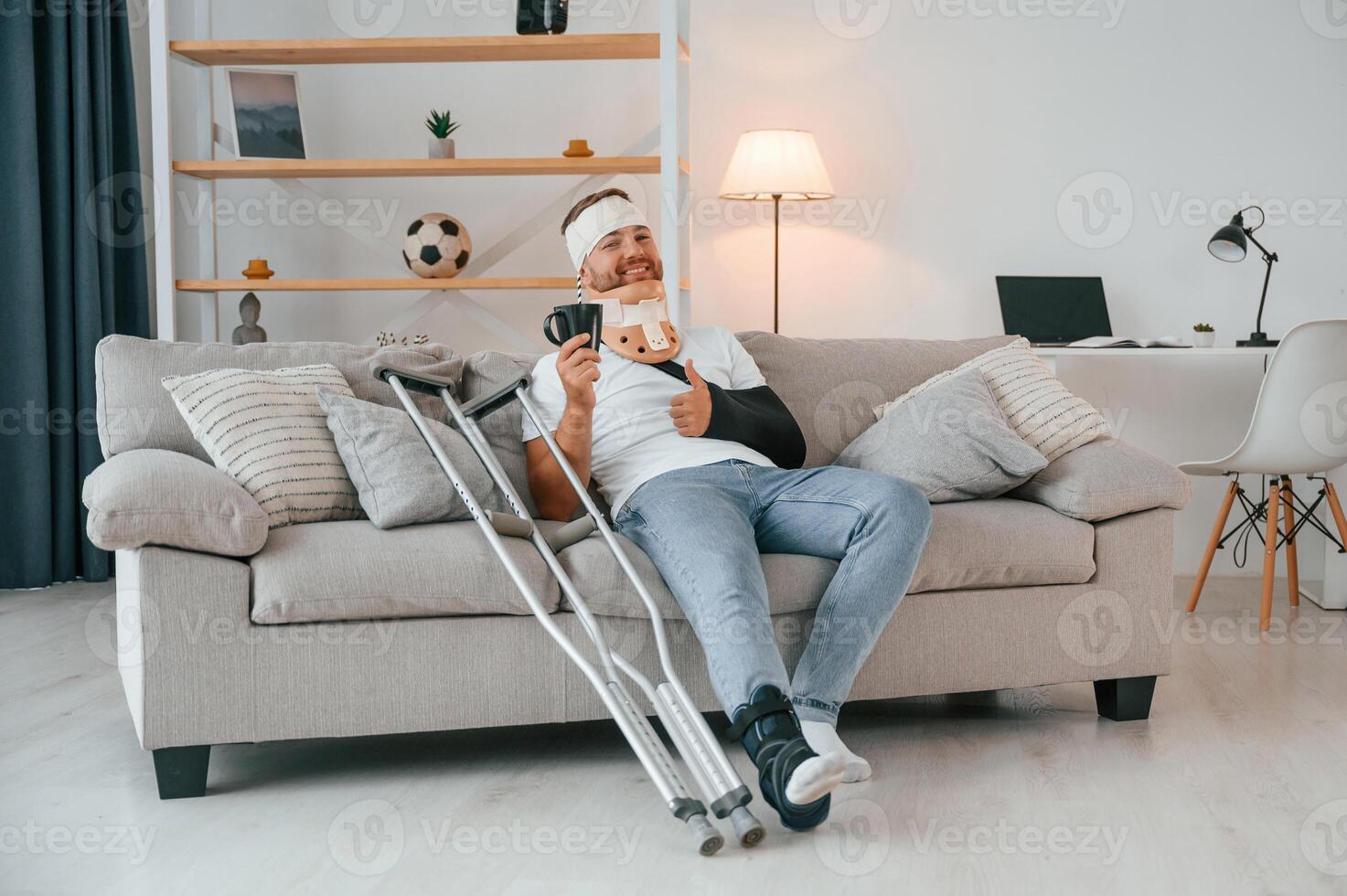 Sitting on the, sofa, showing thumb up, holding cup. Man with crutches is at home indoors. Having injuries photo