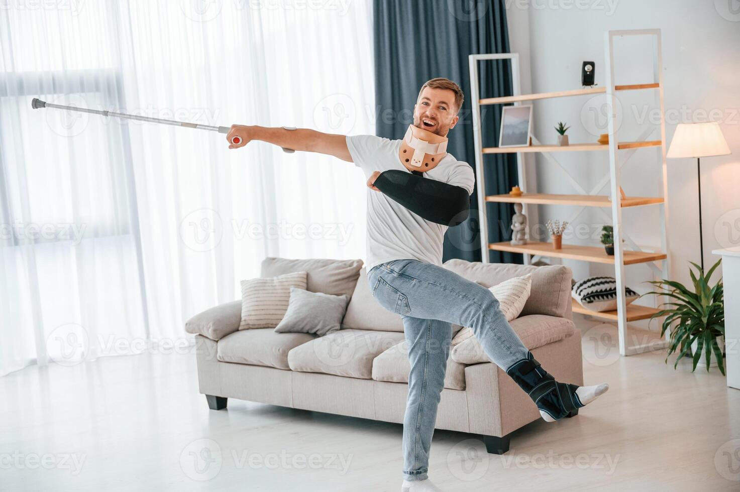 Standing, holding crutch in hand ,and having fun. Man at home indoors. Having injuries photo