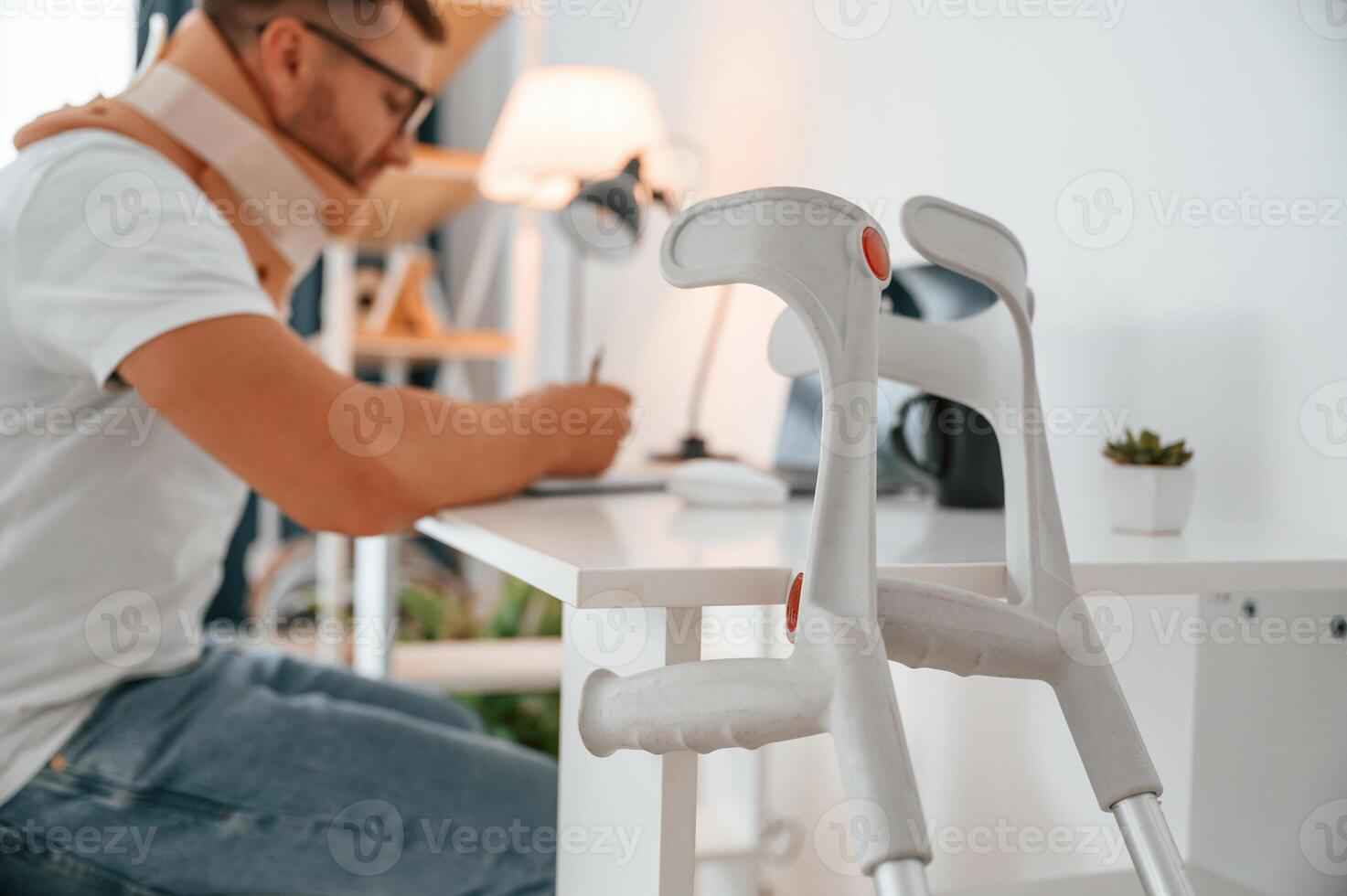 Sitting ,by ,the table and writing. Man with crutches is at home indoors. Having injuries photo