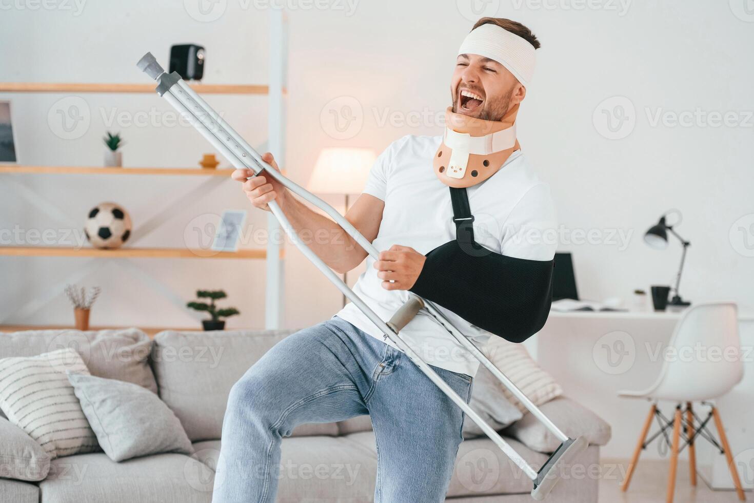 Standing, holding crutch and pretending that he's playing guitar. Having fun. Man is at home indoors. Having injuries photo