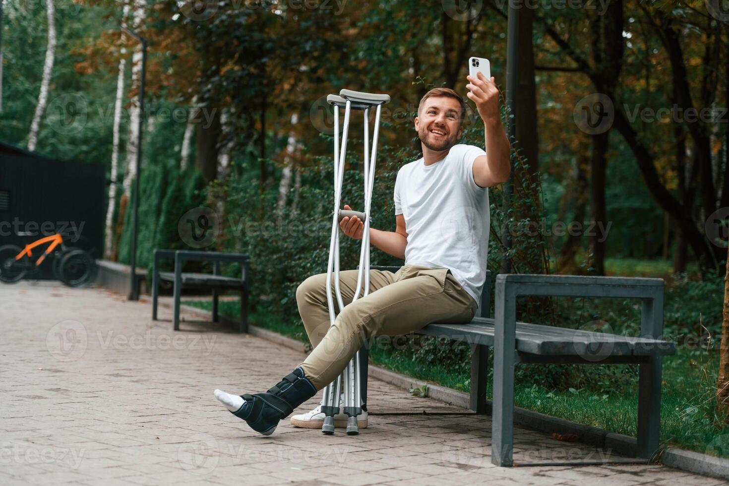Using smartphone to make selfie. Man with ,crutches is in the park outdoors. Having leg injury photo