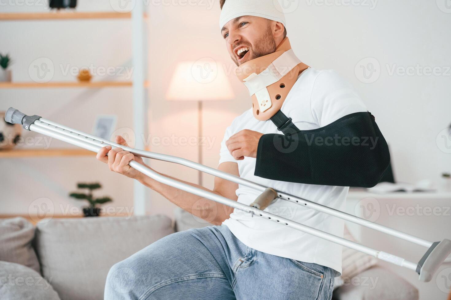 Standing,, holding, crutch and pretending that he's playing guitar. Having fun. Man is at home indoors. Having injuries photo