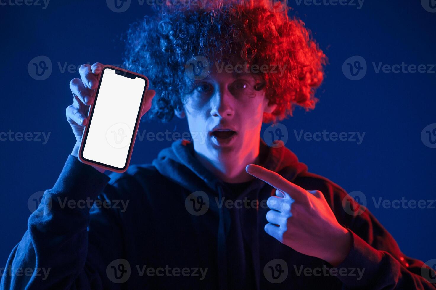 Holding smartphone, and showing it to the camera. Young man with curly hair is indoors illuminated by neon lighting photo