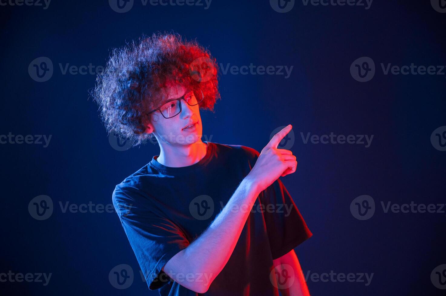 Showing to the right by point finger. Young man with curly hair is indoors illuminated by neon lighting photo