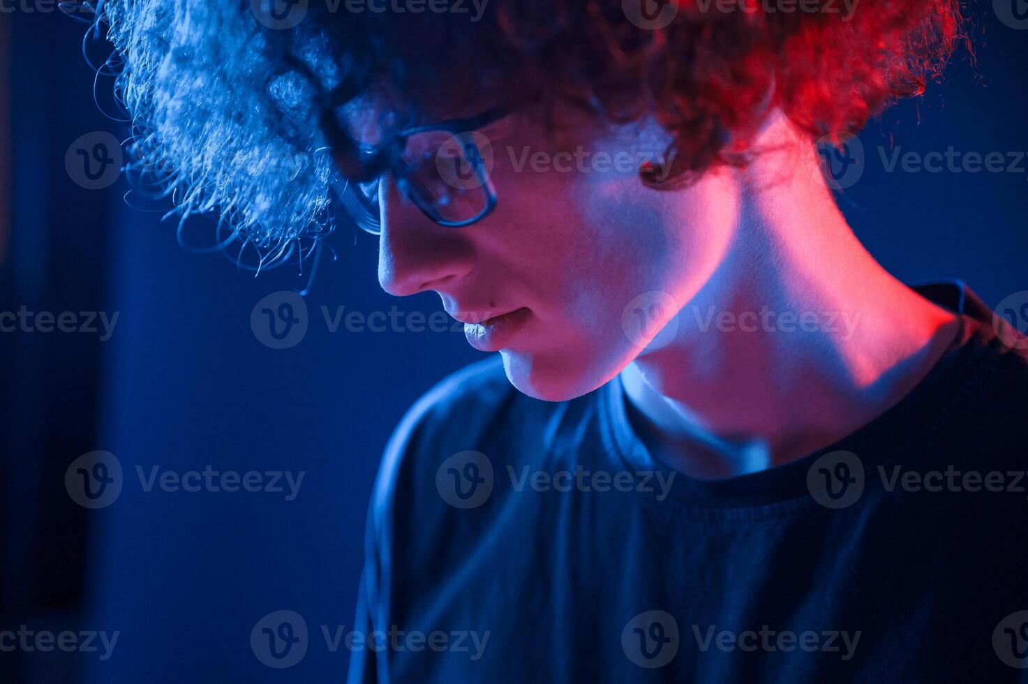 Looking down. Young man with curly hair is indoors illuminated by neon lighting photo