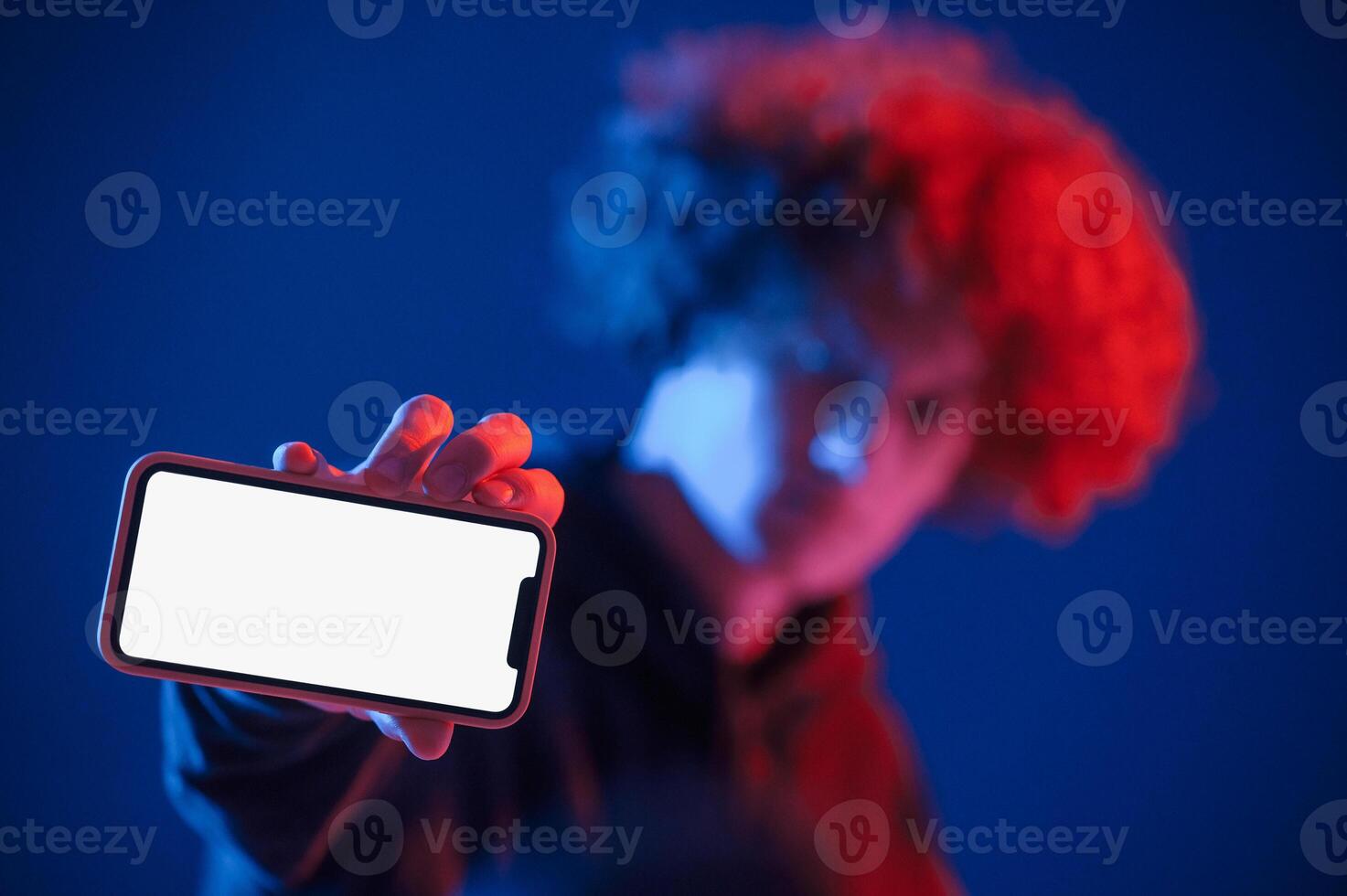 Holding smartphone and showing it to the camera. Young man with curly hair is indoors illuminated by neon lighting photo