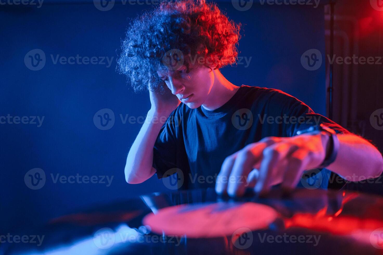 Dj is playing music from vinyl.. Young man with curly hair is indoors illuminated by neon lighting photo
