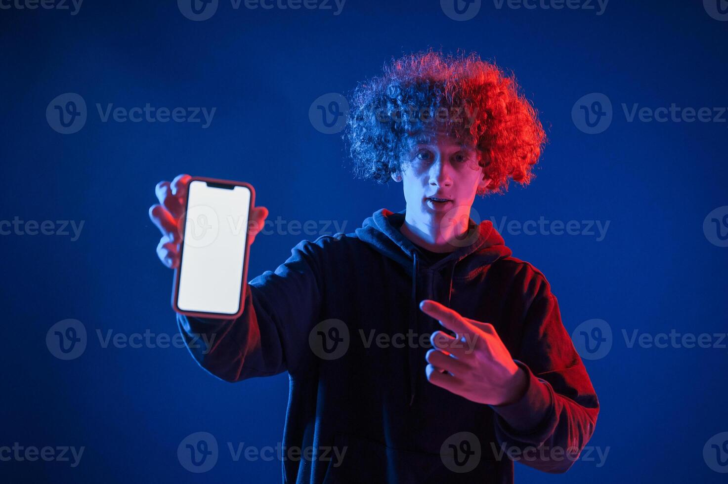 Holding smartphone and showing, it to the camera. Young man with curly hair is indoors illuminated by neon lighting photo