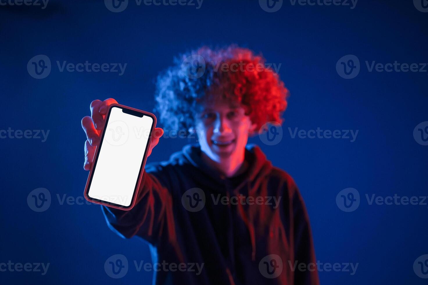 Holding smartphone ,and showing it to the camera. Young man with curly hair is indoors illuminated by neon lighting photo