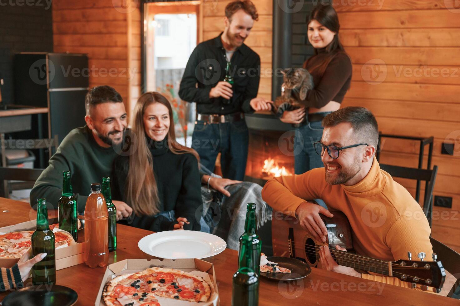 Man, in glasses is playing guitar. Group of friends is having good weekend indoors in the wooden building together photo