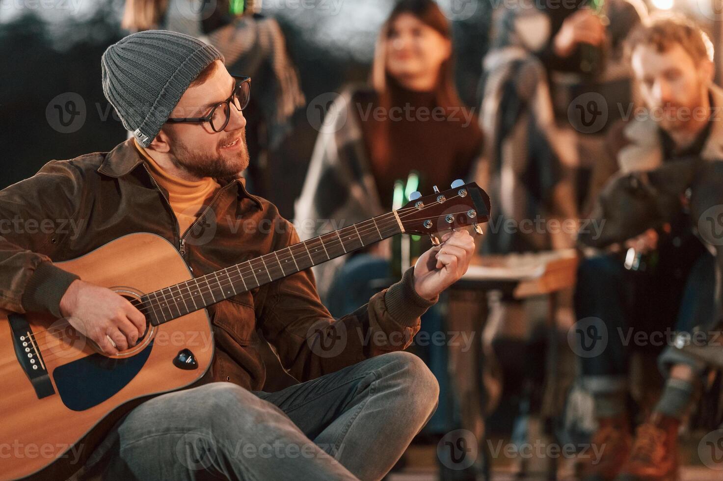 Man in ,hat is sitting ,and playing guitar. Group of people is spending time together on the backyard at evening time photo