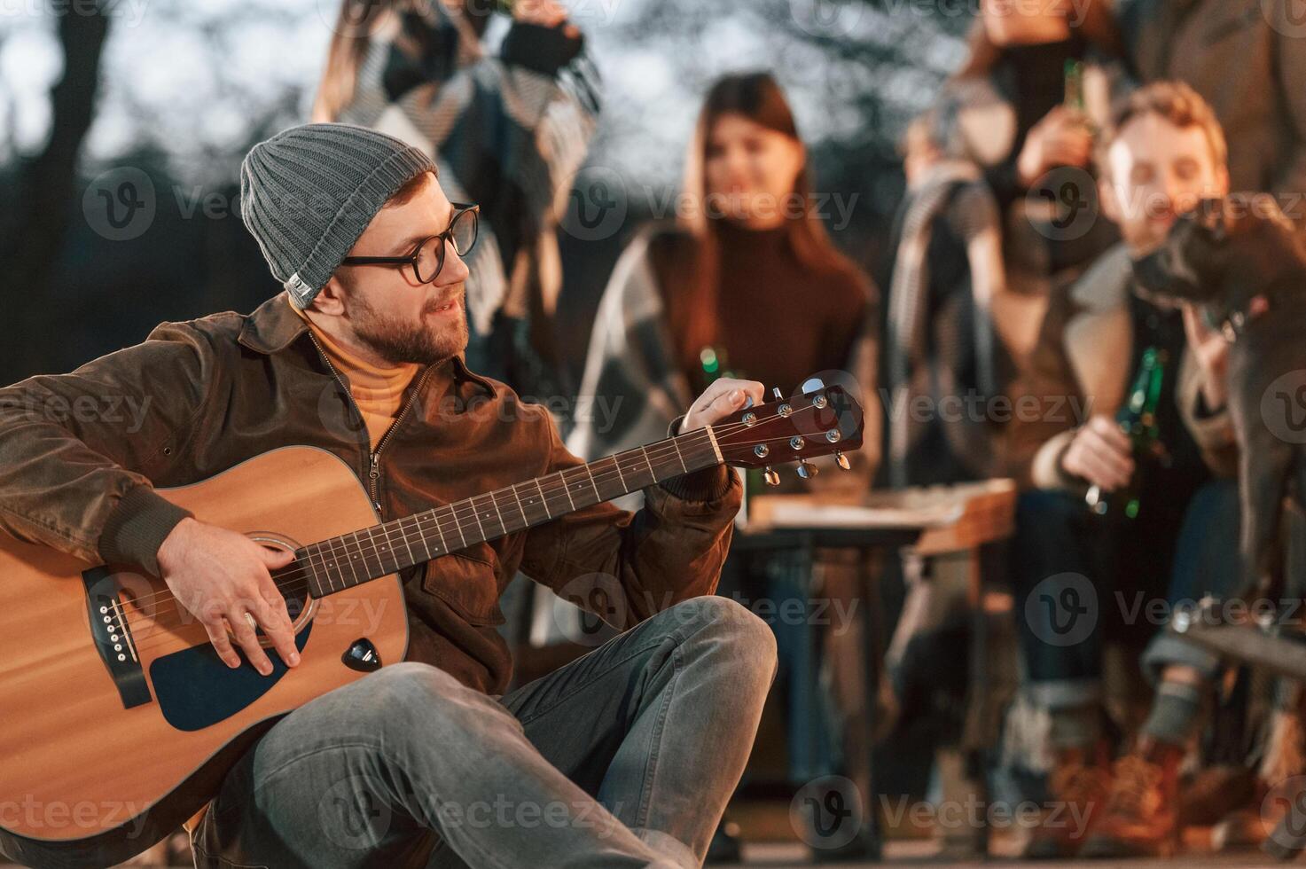Man in hat is sitting and playing guitar. Group of people is spending time together on the backyard at evening time photo