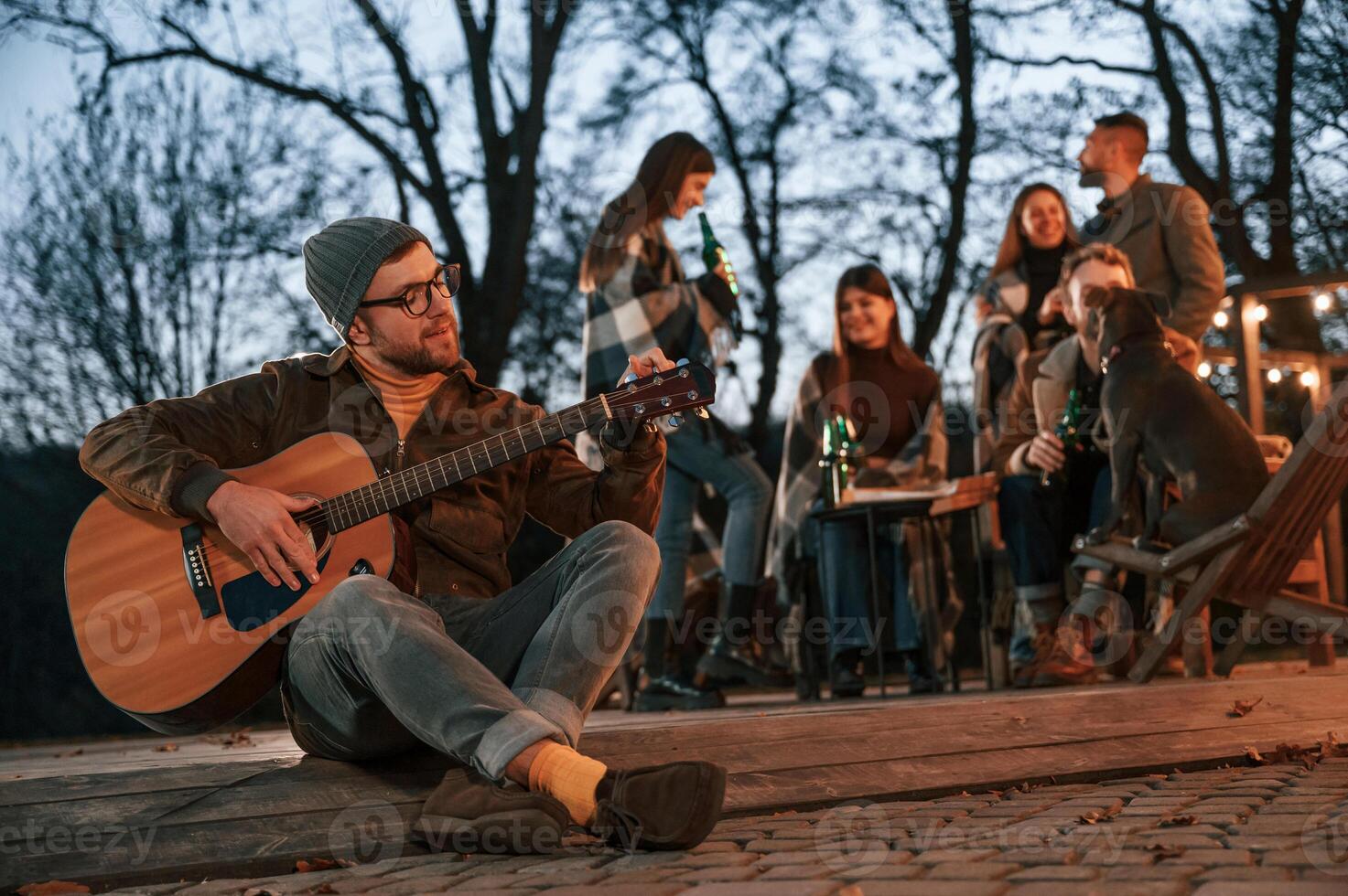 Man with ,guitar is sitting, in front of his friends. Group of people is spending time together on the backyard photo