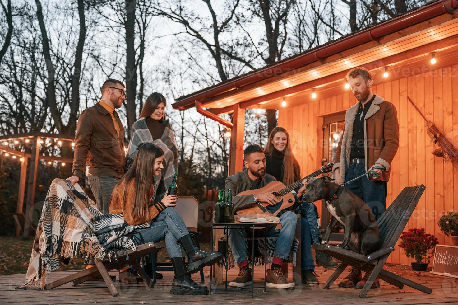 Man playing guitar, dog ,is sitting on the chair. Group of people is spending time together on the backyard at evening time photo