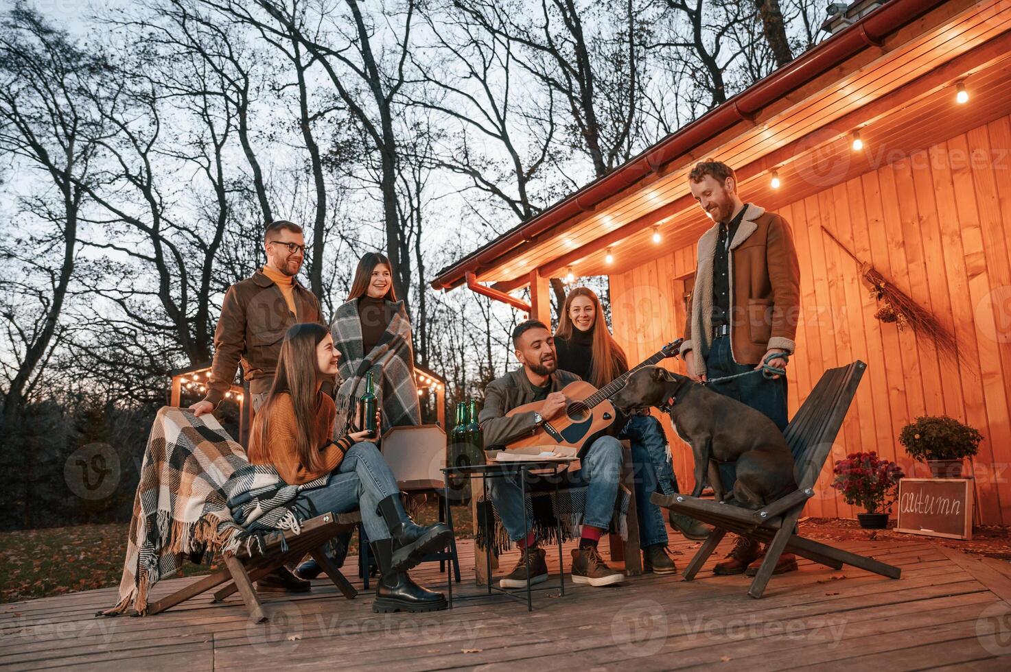 Man playing guitar,, dog is sitting on the chair. Group of people is spending time together on the backyard at evening time photo