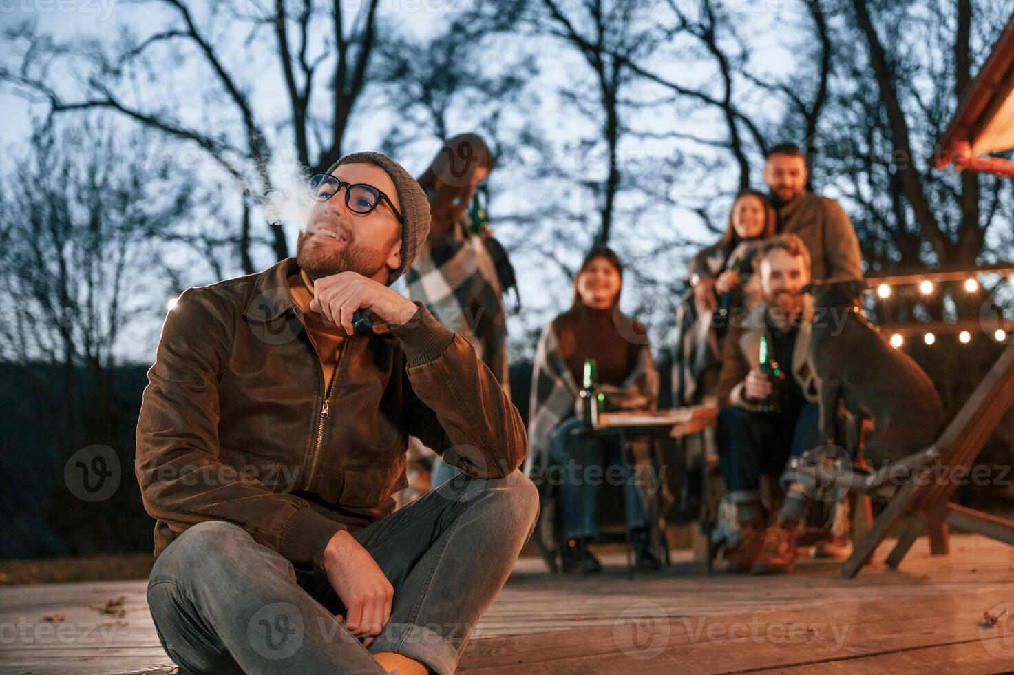 Man is, smoking. Group of people is spending time together on the backyard at evening time photo