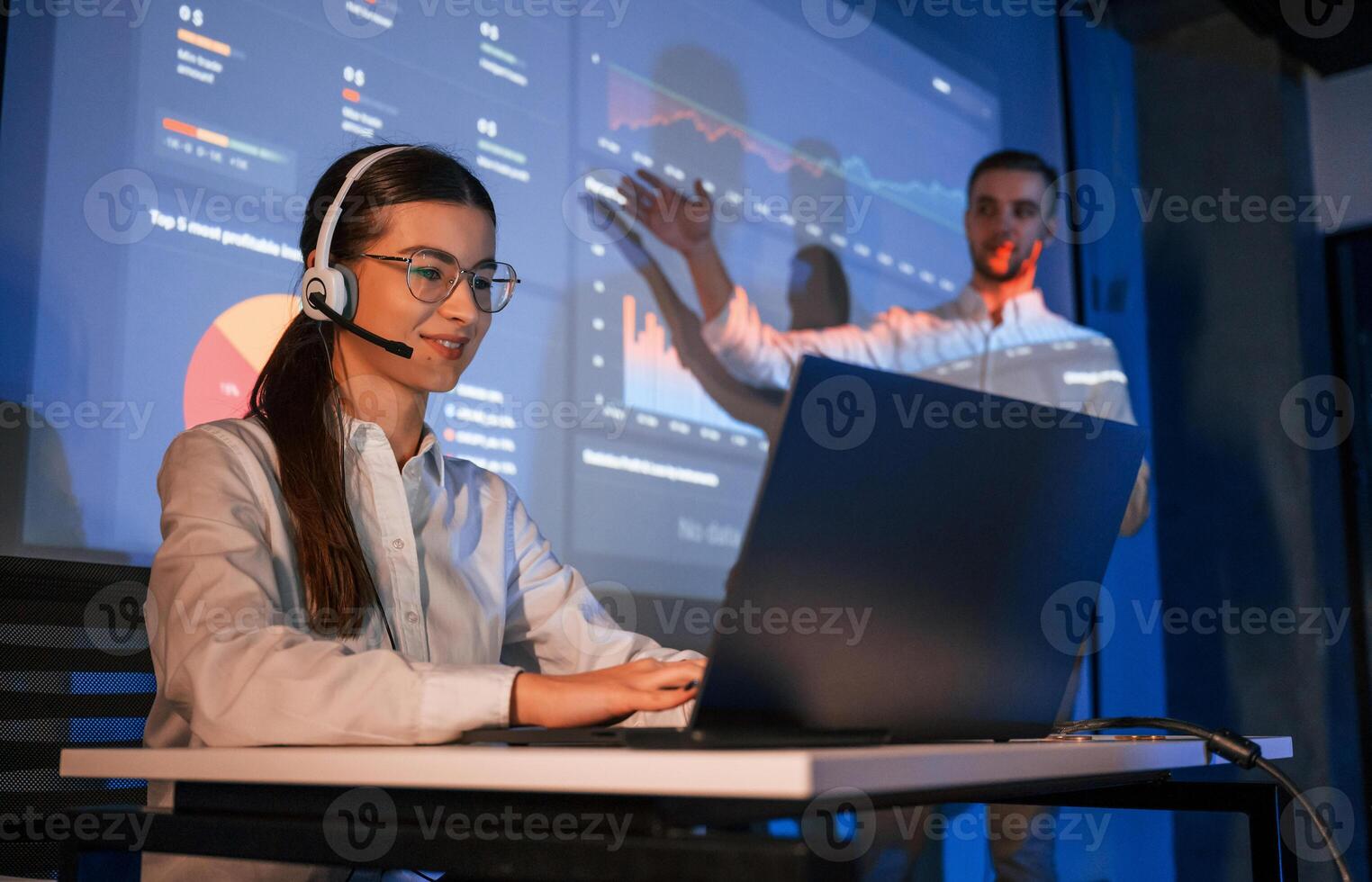 Stock broker is sitting and ,working by laptop. Man is standing near her, showing data on projector photo