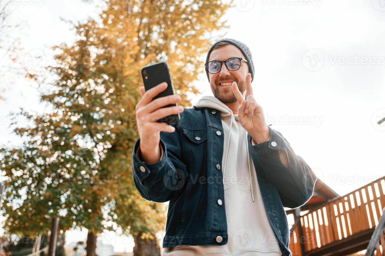 call by using smartphone, showing two fingers. Beautiful man in warm clothes is outdoors in the park photo