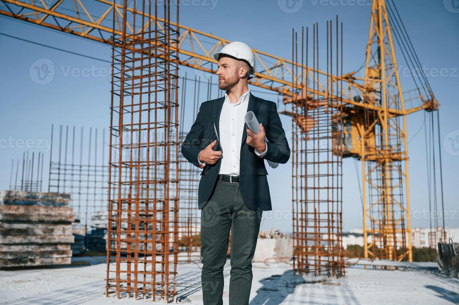 In hard, hat, holding plan in hands. Businessman in formal clothes is on the construction site at daytime photo