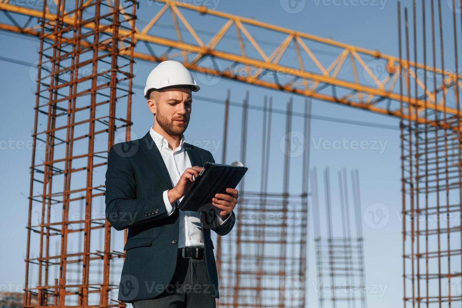 Holding paper with, plan and tablet. Businessman in formal clothes is on the construction site at daytime photo