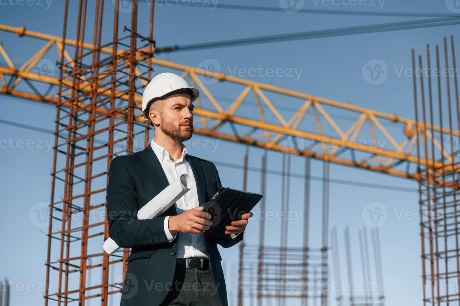 In white, hard hat, with plan and tablet in hands. Businessman in formal clothes is on the construction site at daytime photo