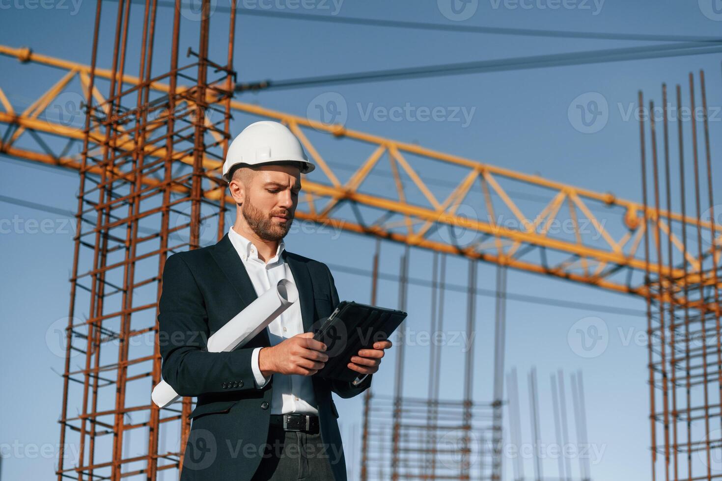 In ,white, hard hat, with plan and tablet in hands. Businessman in formal clothes is on the construction site at daytime photo