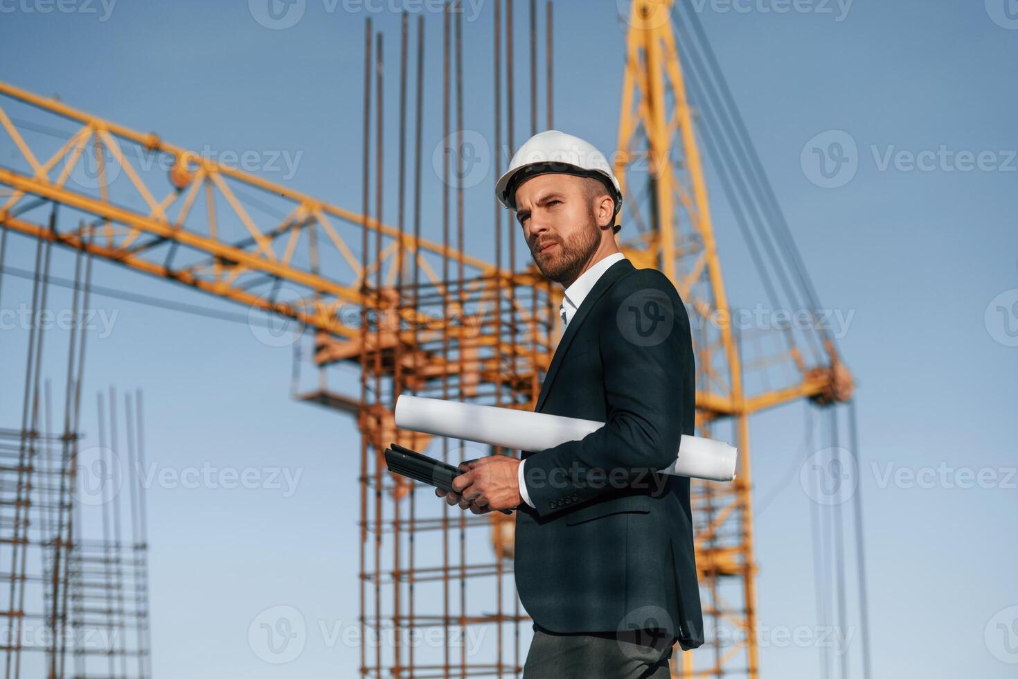 Side view. Walking and holding ,paper with, plan and digital tablet. Businessman in formal clothes is on the construction site photo