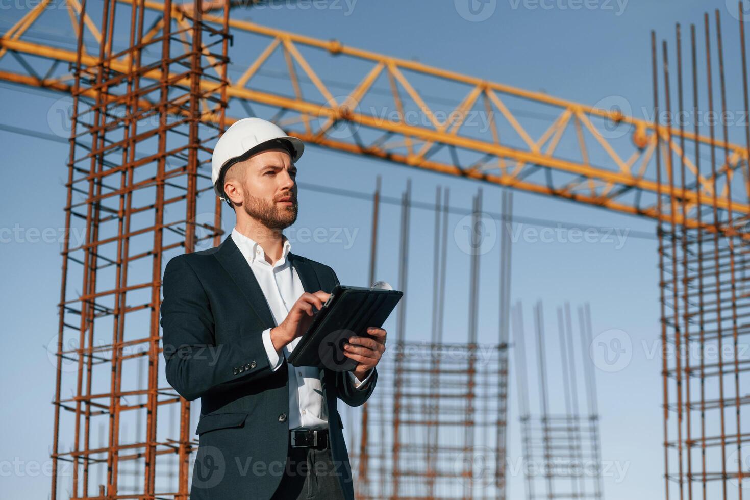 In white hard hat,. with plan and tablet in hands. Businessman in formal clothes is on the construction site at daytime photo