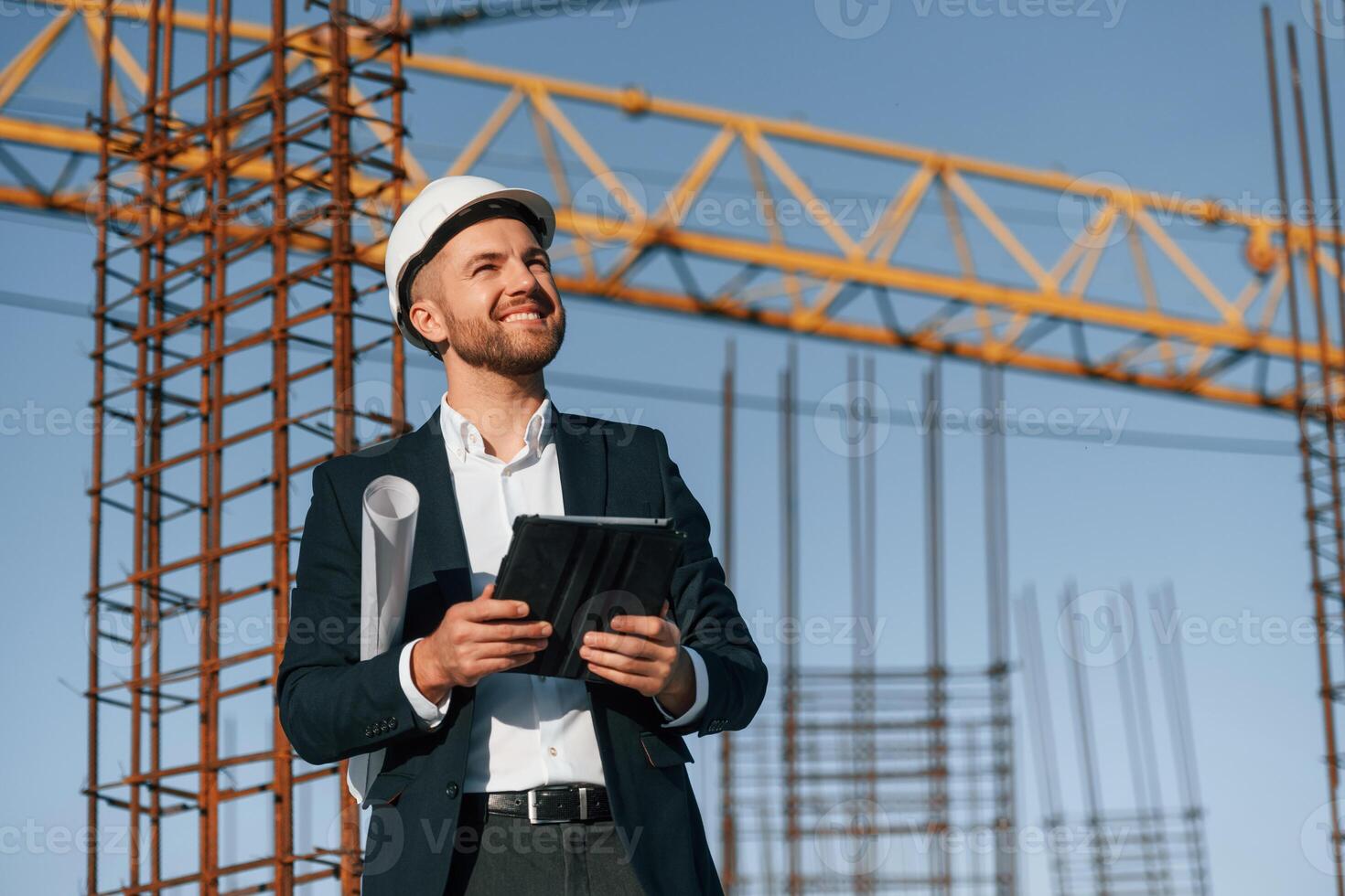 In white ,hard hat, with ,plan and tablet in hands. Businessman in formal clothes is on the construction site at daytime photo