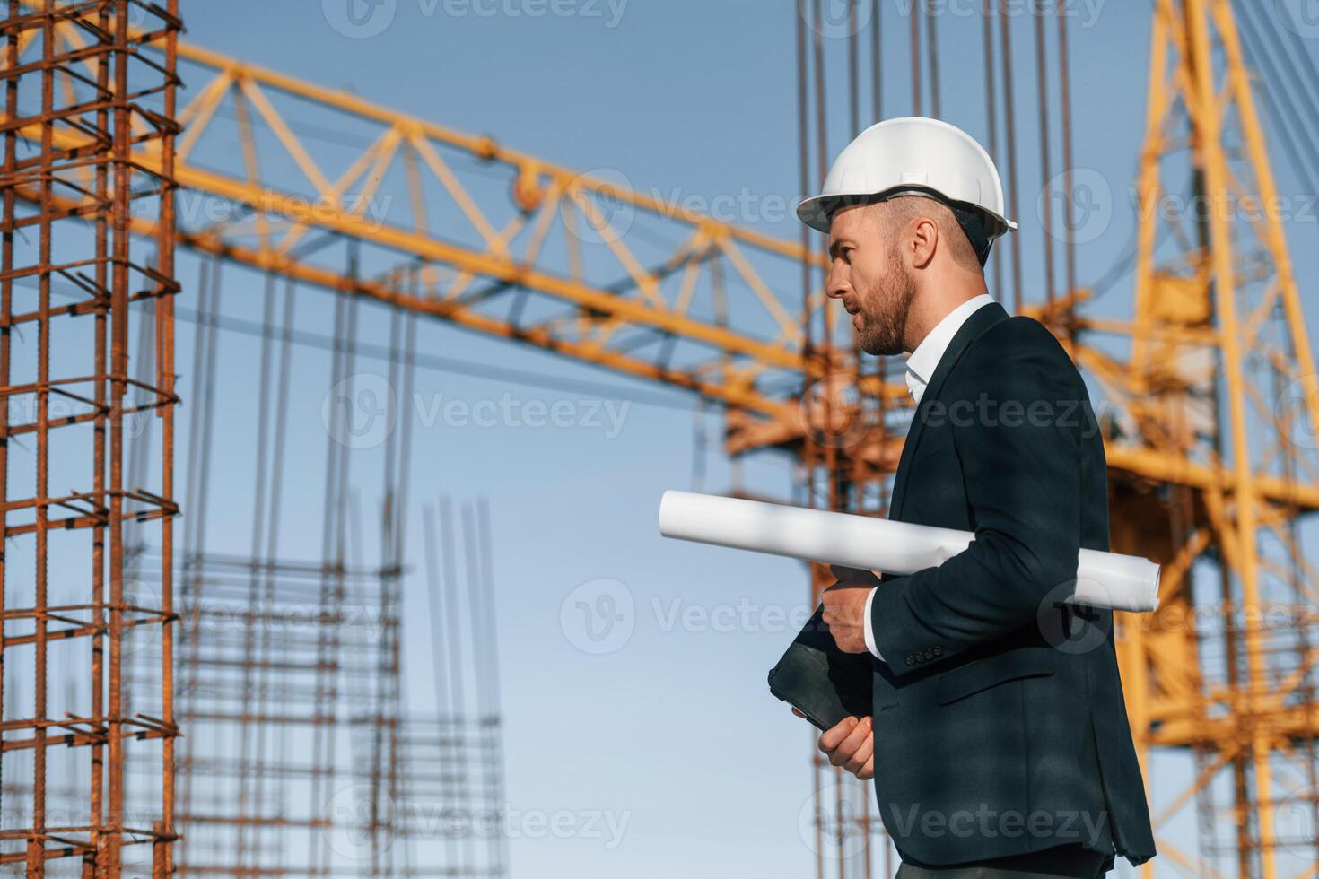 Side view. Walking, and, holding paper with plan and digital tablet. Businessman in formal clothes is on the construction site photo