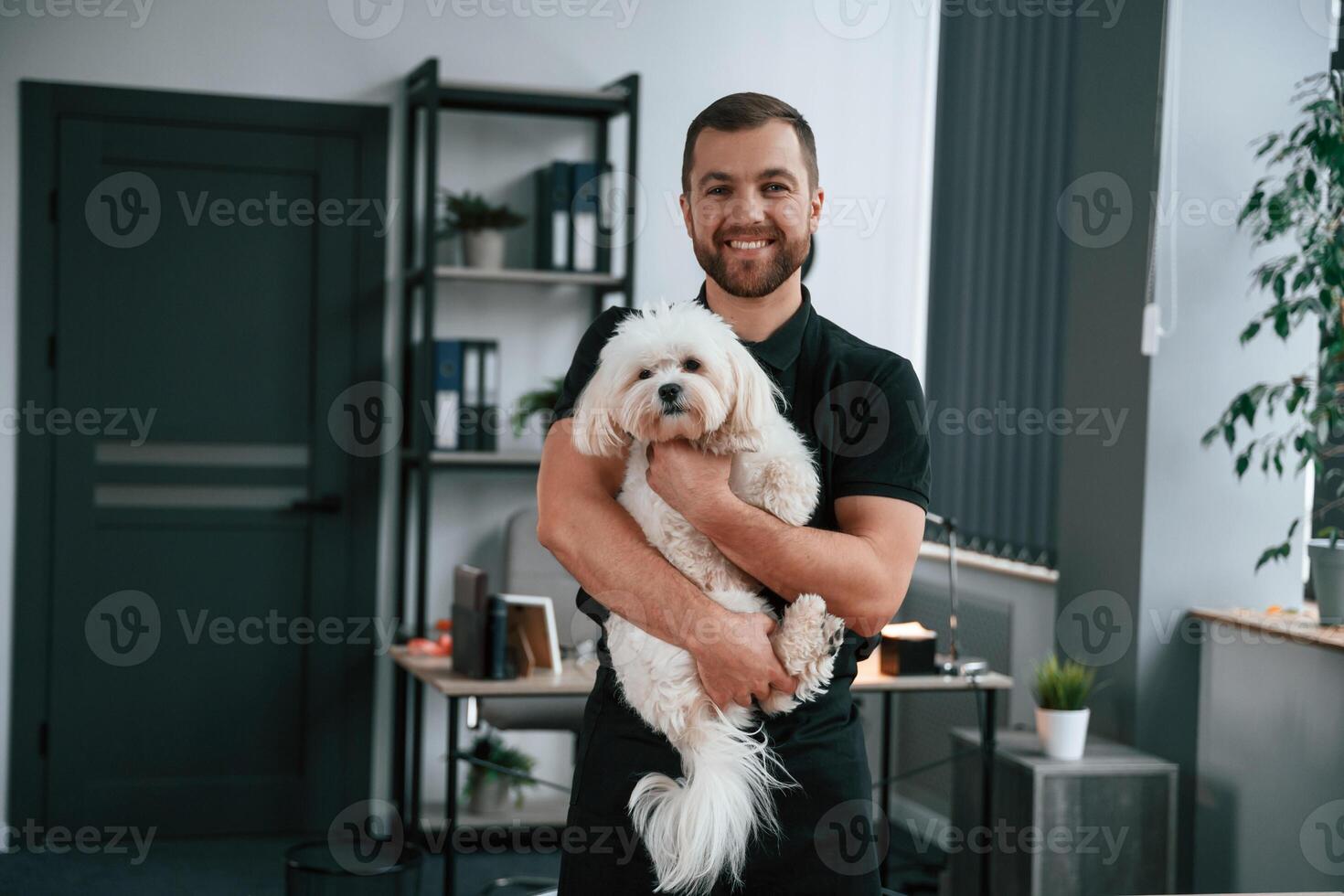 Man is smiling.. Groomer, is with, Maltese dog indoors. Man is in the black uniform photo