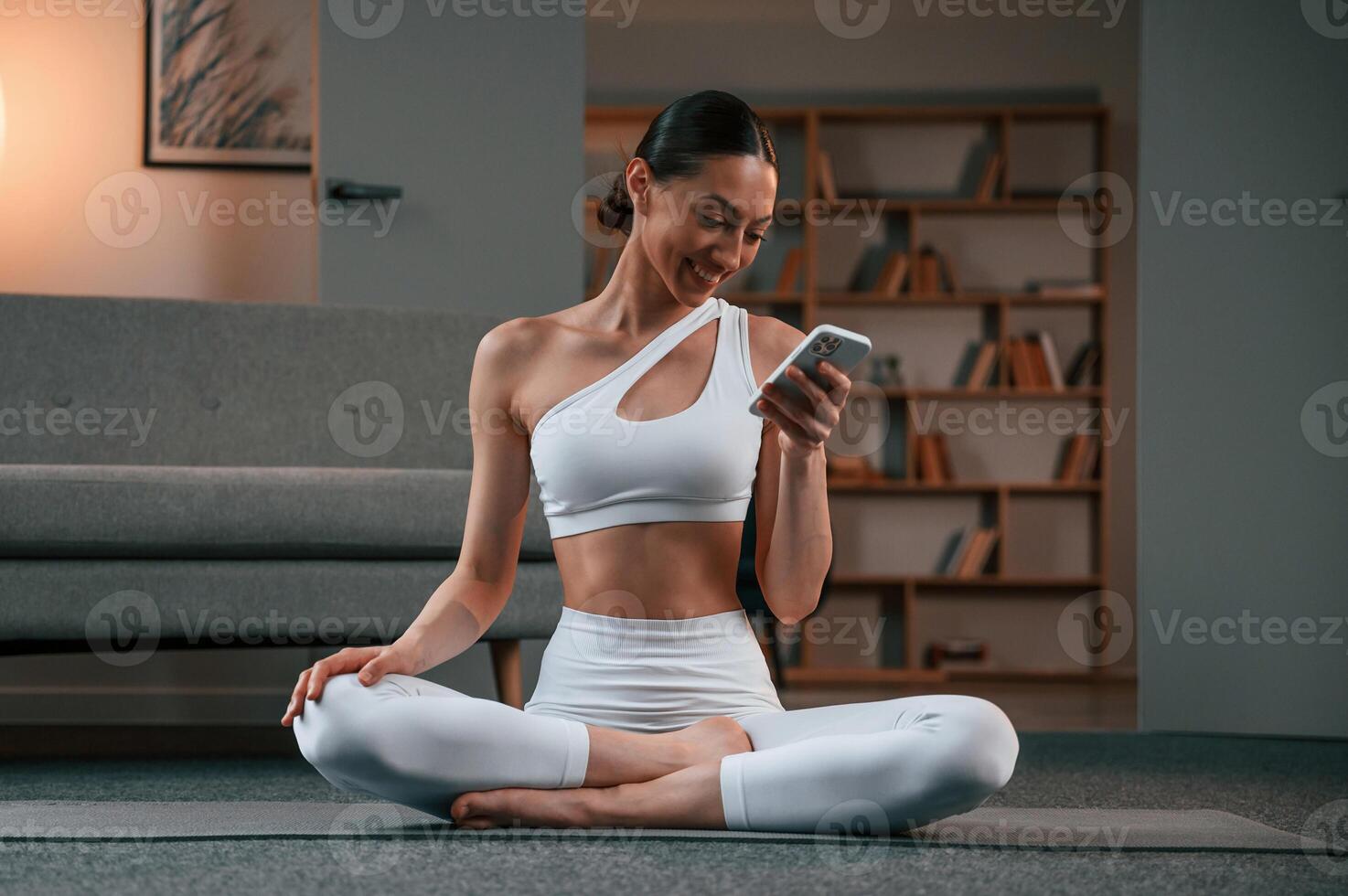Holding smartphone. Young woman with, slim body type in white sportswear is indoors doing yoga photo
