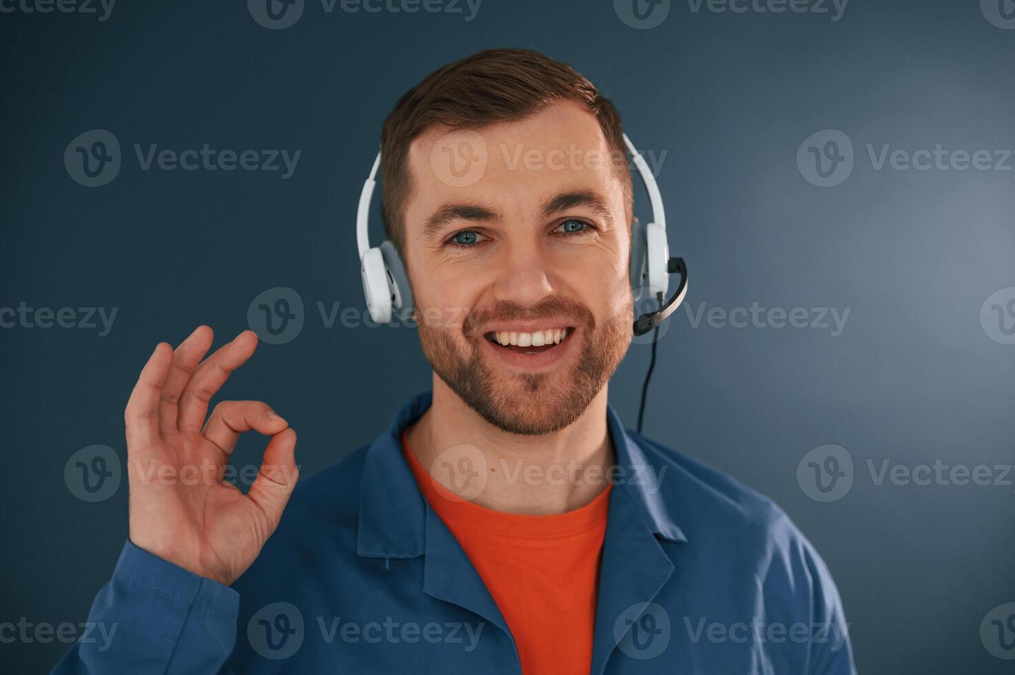 Showing fine, okay, good, sign., Smiling. In headphones. Handsome man is in the studio against blue background photo