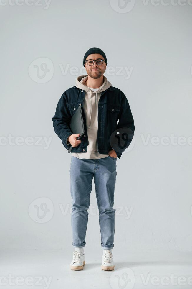 IT specialist. in hat and glasses standing with laptop. Handsome man is in the studio against white background photo