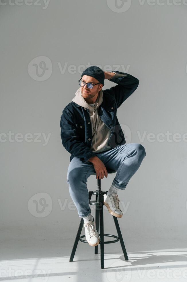 Model, in hat and glasses is sitting on a chair. Handsome man is in the studio against white background photo