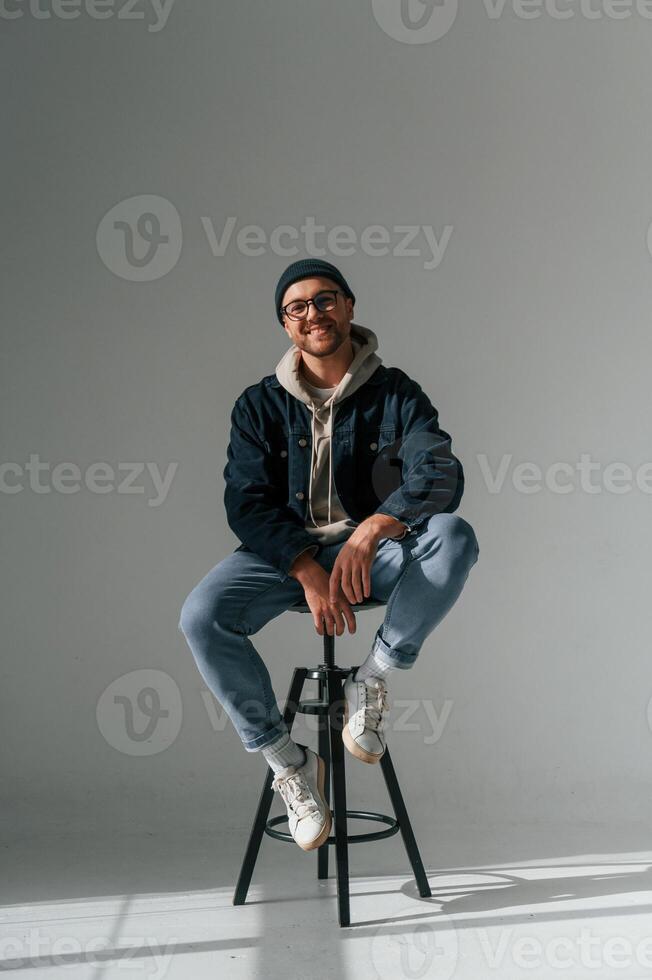Model in hat and glasses is sitting on a chair. Handsome man is in the studio against white background photo