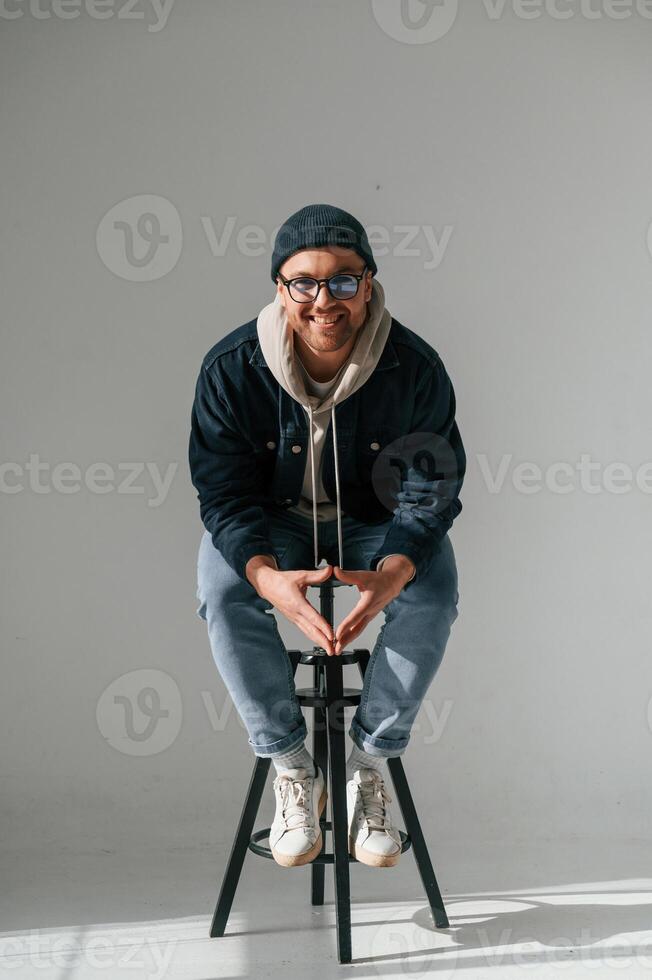 Model in hat and glasses is sitting on a, chair. Handsome man is in the studio against white background photo