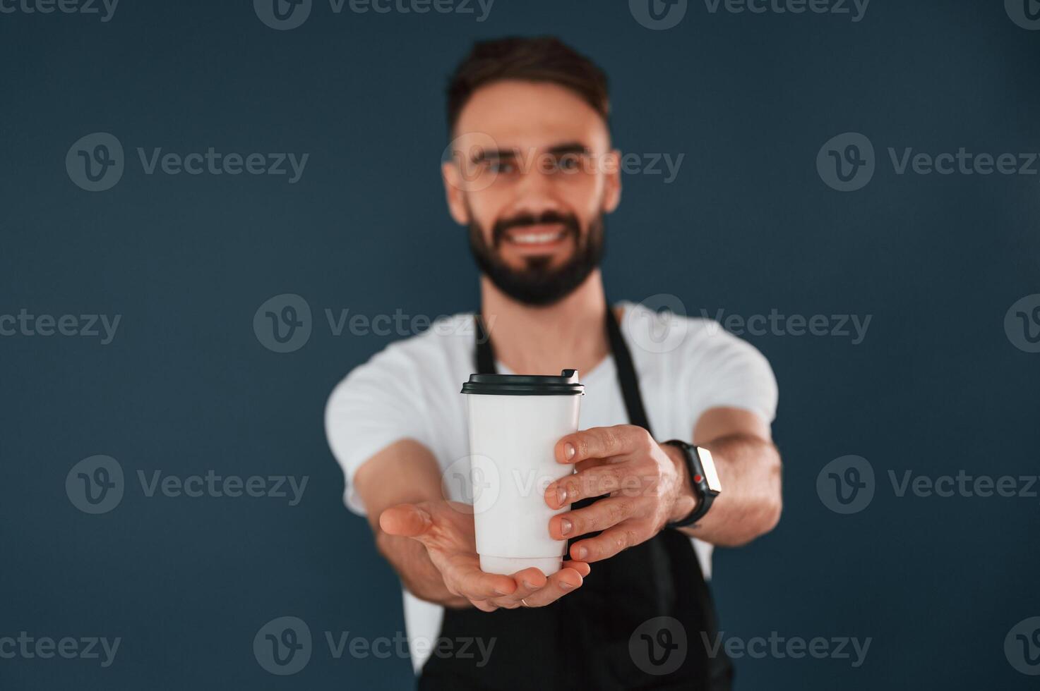 Giving the ,cup ,of coffee. Cafe worker is in apron. Handsome man is in the studio against blue background photo
