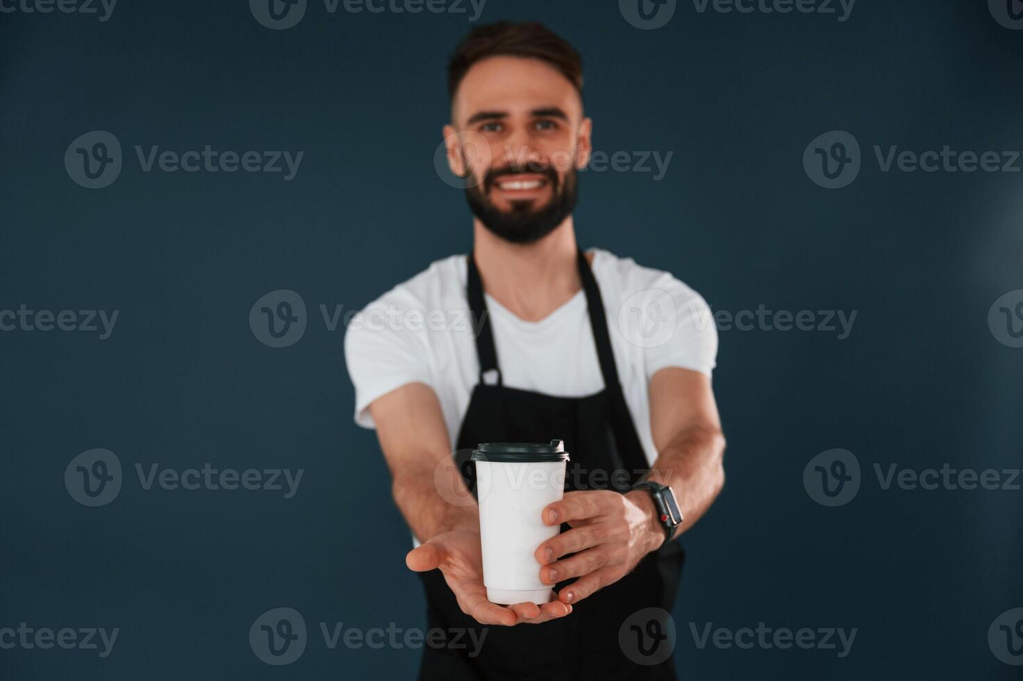 Giving the cup of coffee.. Cafe worker is in apron. Handsome man is in the studio against blue background photo