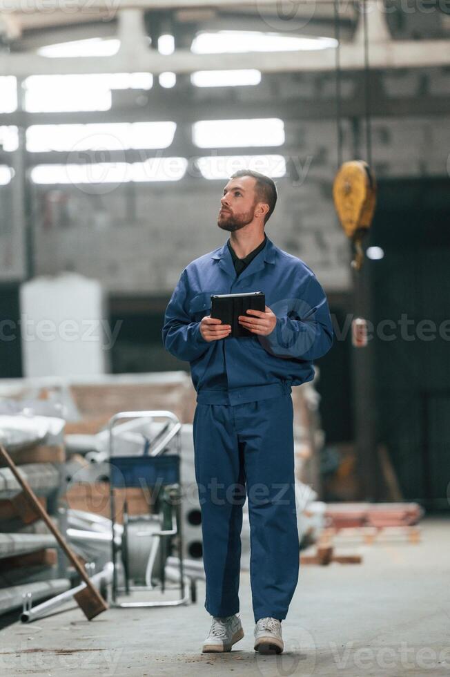 With tablet in hands. Factory ,worker in blue uniform is indoors photo